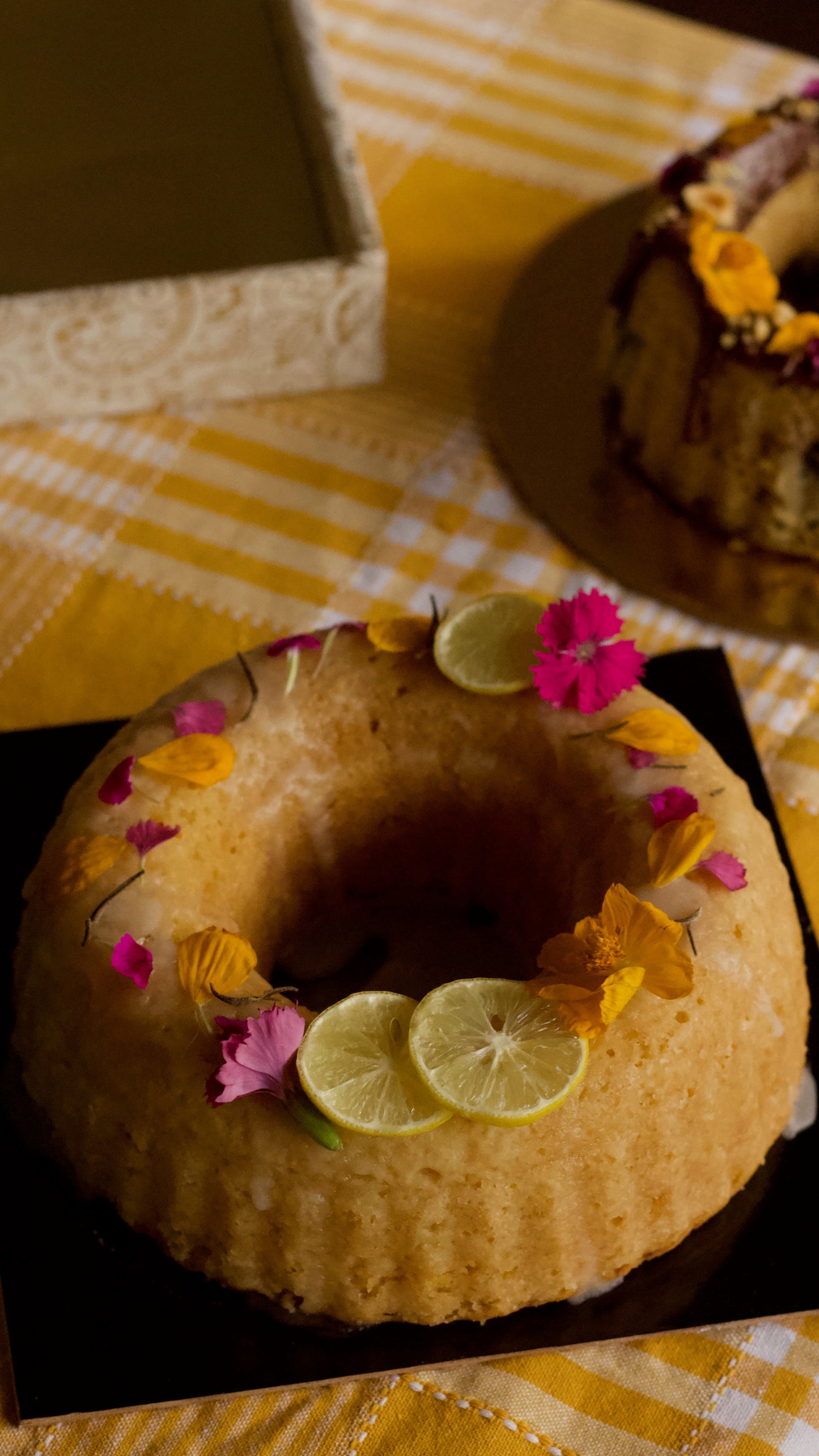 A ring-shaped bundt cake decorated with lemon slices and edible flowers, presented on a black square plate over a yellow-and-white checkered tablecloth. A matching decorative box is blurred in the background.