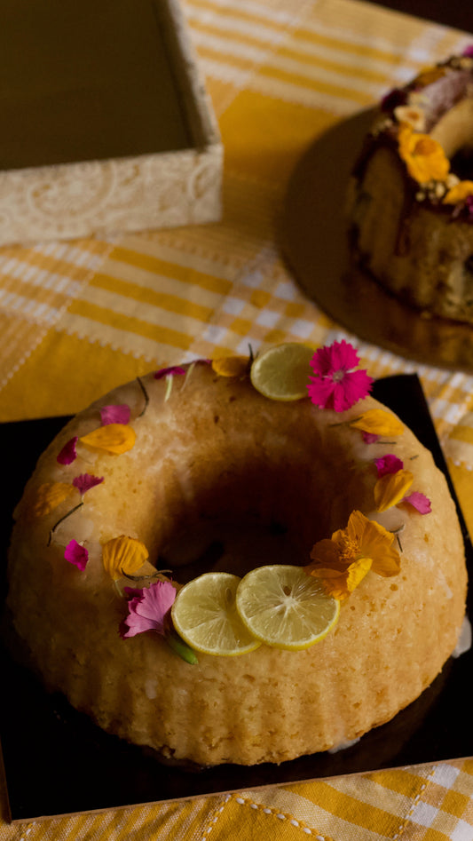 A ring-shaped bundt cake decorated with lemon slices and edible flowers, presented on a black square plate over a yellow-and-white checkered tablecloth. A matching decorative box is blurred in the background.