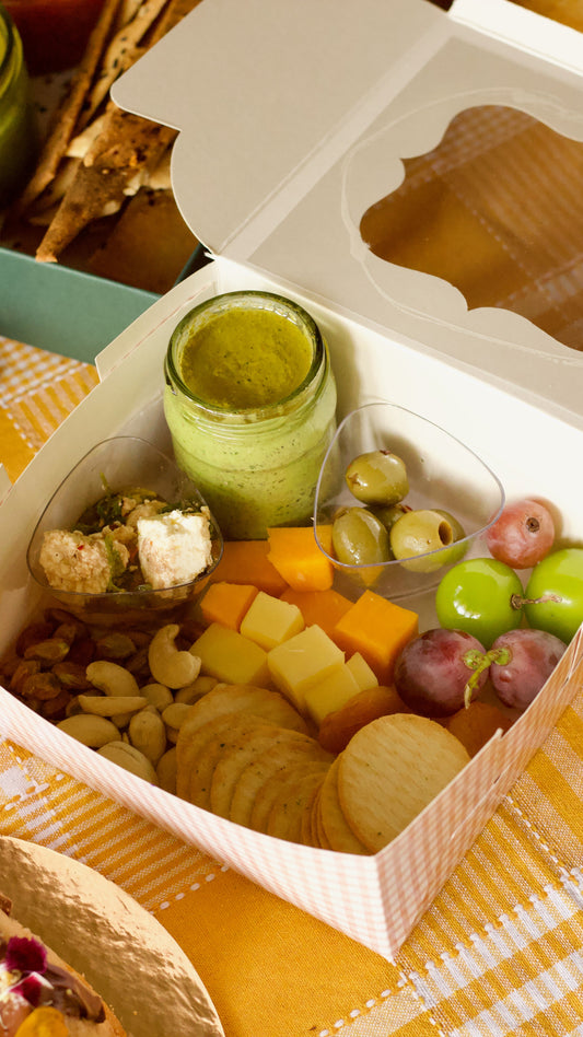 A gourmet charcuterie box filled with crackers, cheese cubes, grapes, olives, nuts, and a small jar of green dip, presented in a white paper box on a yellow-and-white checkered tablecloth.