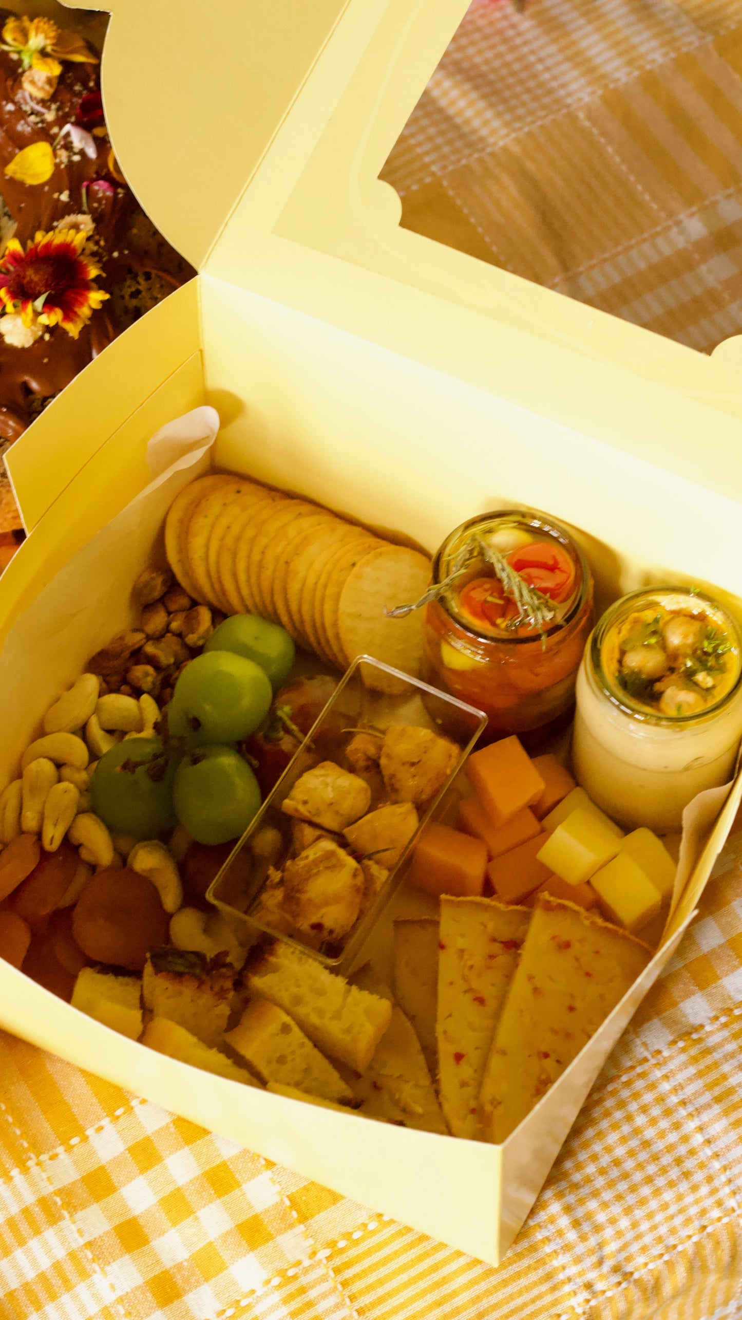 A gourmet charcuterie board box filled with crackers, cheese cubes, nuts, green olives, dips, and small jars, presented in a yellow paper box on a yellow-and-white checkered blanket.