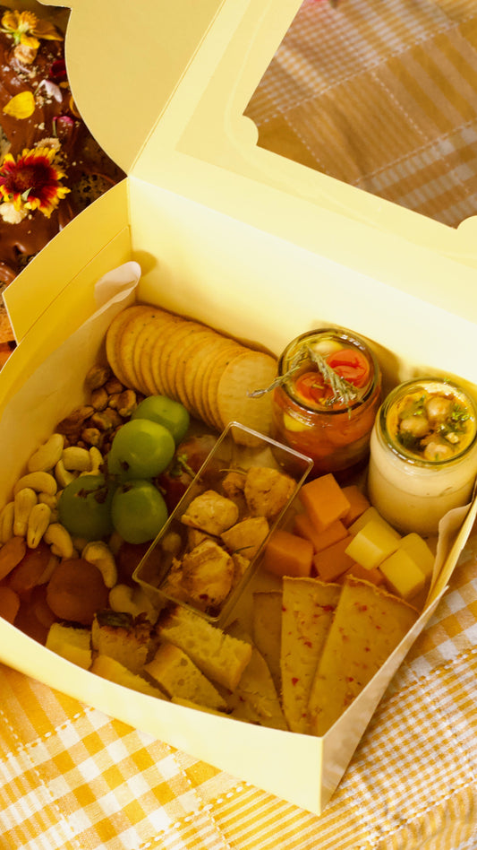 A gourmet charcuterie board box filled with crackers, cheese cubes, nuts, green olives, dips, and small jars, presented in a yellow paper box on a yellow-and-white checkered blanket.