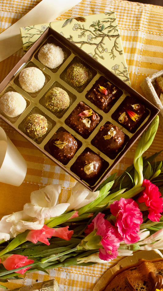 An open box of assorted  vegan gluten free refined sugar free sweet bites laddoos arranged on a yellow-and-white checkered tablecloth with flowers in the foreground. 
